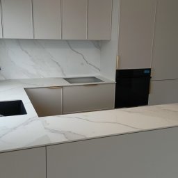 Stablo Meble - Jasna kuchnia z marmurowym blatem i złotymi uchwytami. Nowoczesny design z wyspą kuchenną i wbudowanym sprzętem AGD. Minimalistyczne szafki wiszące i podłogowe.