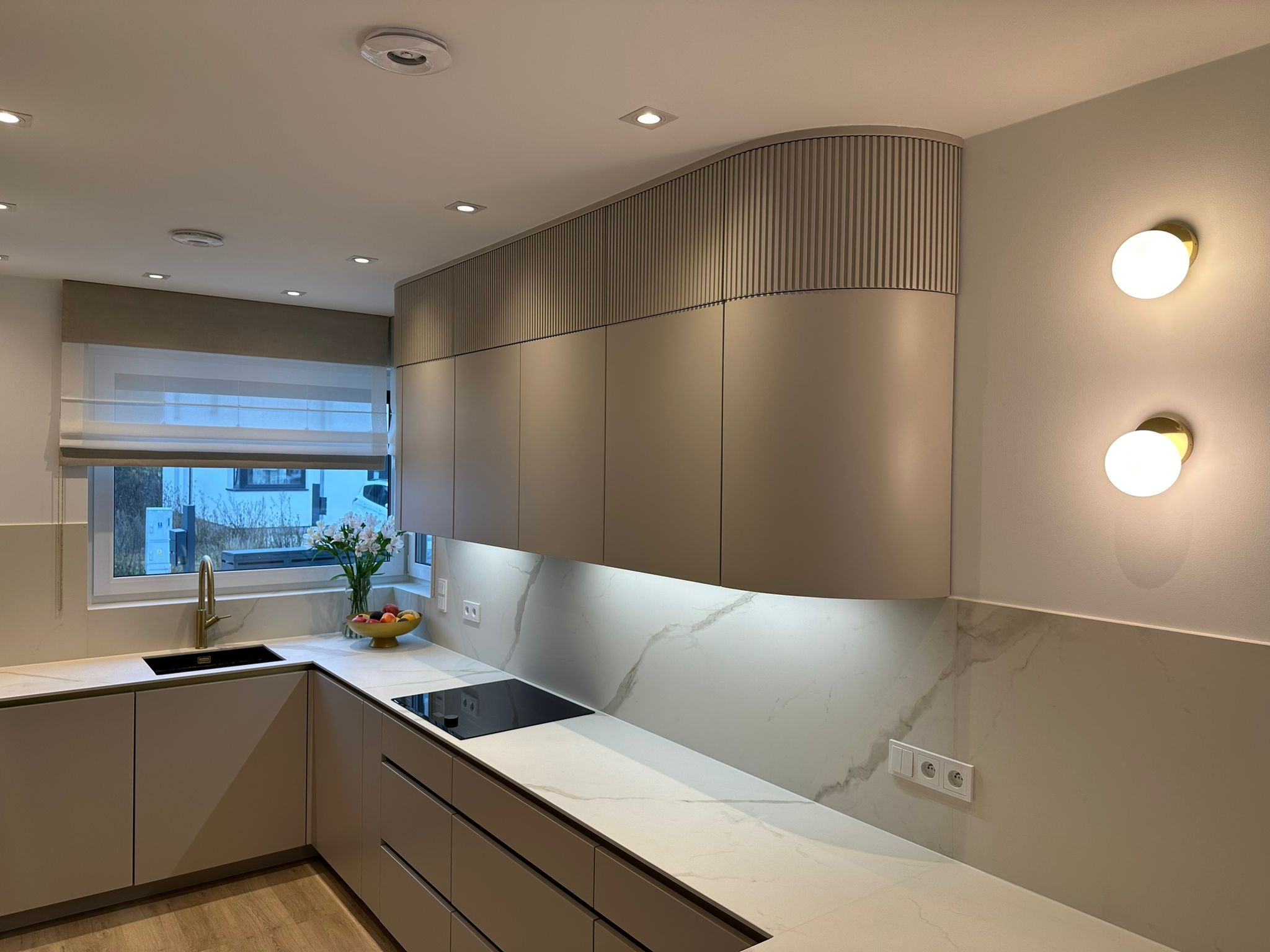 Nowoczesna kuchnia z beżowymi szafkami, frezowanym wykończeniem górnych szafek, białym blatem z marmurkowym wzorem i złotą baterią przy zlewie pod oknem.