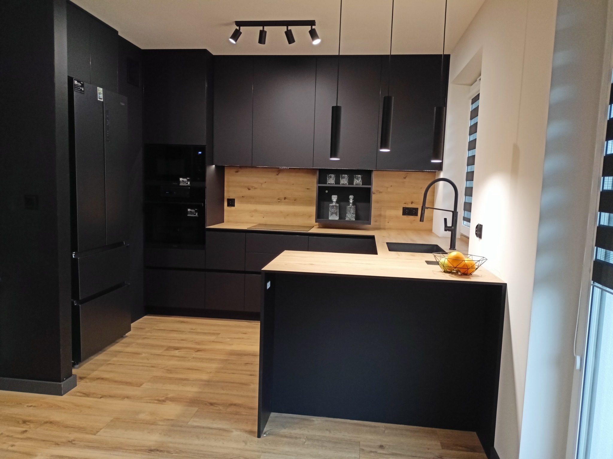 Nowoczesna, czarna kuchnia z drewnianymi akcentami, oświetleniem punktowym i wyspą z owocami w metalowym koszyku, widok na okno z żaluzjami.