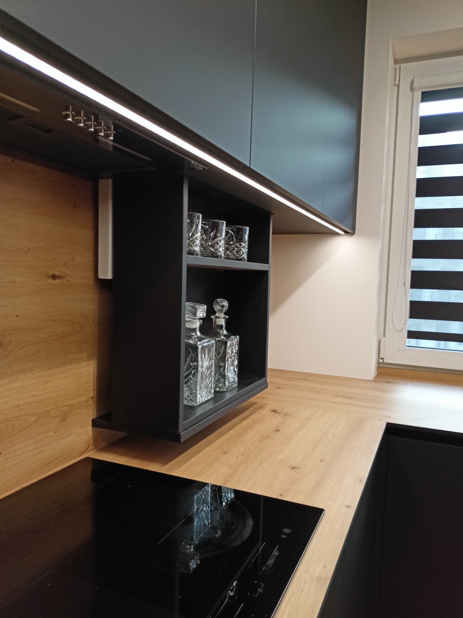 Nowoczesna kuchnia z czarnymi szafkami i blatem drewnopodobnym, oświetlenie LED pod szafkami, półka z szklankami i karafkami, płyta indukcyjna w blacie.