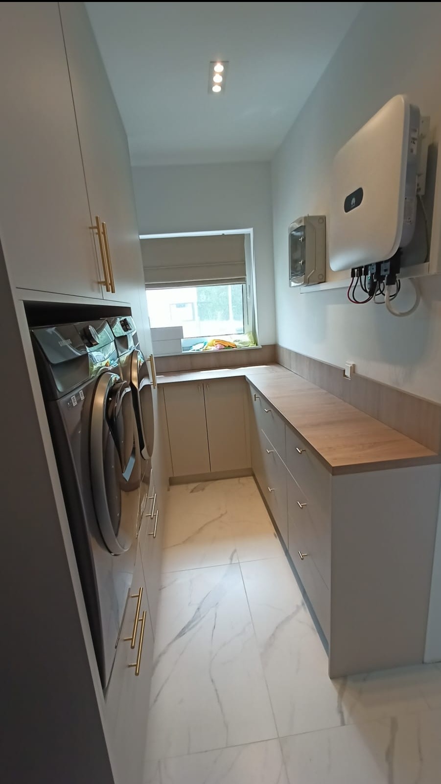 Funkcjonalna pralnia w Szczecinie z zabudową meblową na wymiar, zawierająca pralki, szafki z drewnianym blatem i białą marmurową podłogą, oświetlona trzema punktami świetlnymi.