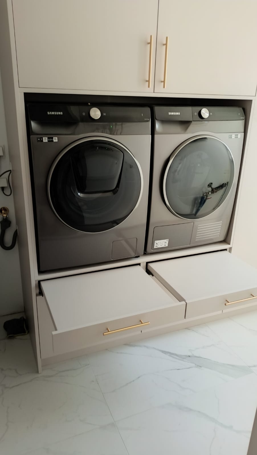 Szaro-beżowa zabudowa z pralką i suszarką Samsung, szuflady z poziomymi, złotymi uchwytami, marmurowa podłoga.