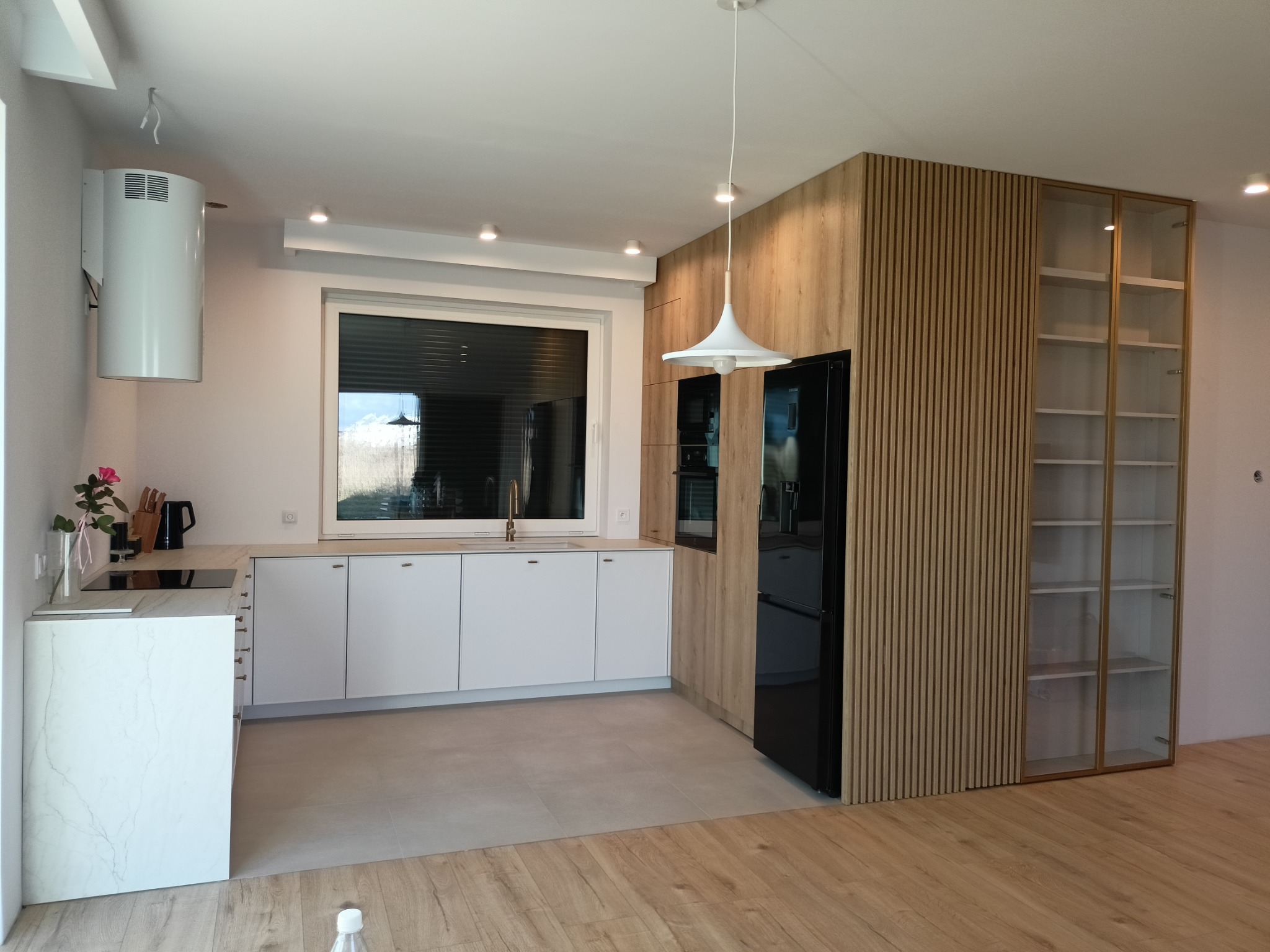 Nowoczesna kuchnia z białymi frontami, drewnianymi panelami ściennymi i czarną lodówką, oświetlona wiszącą lampą w kształcie kapelusza.