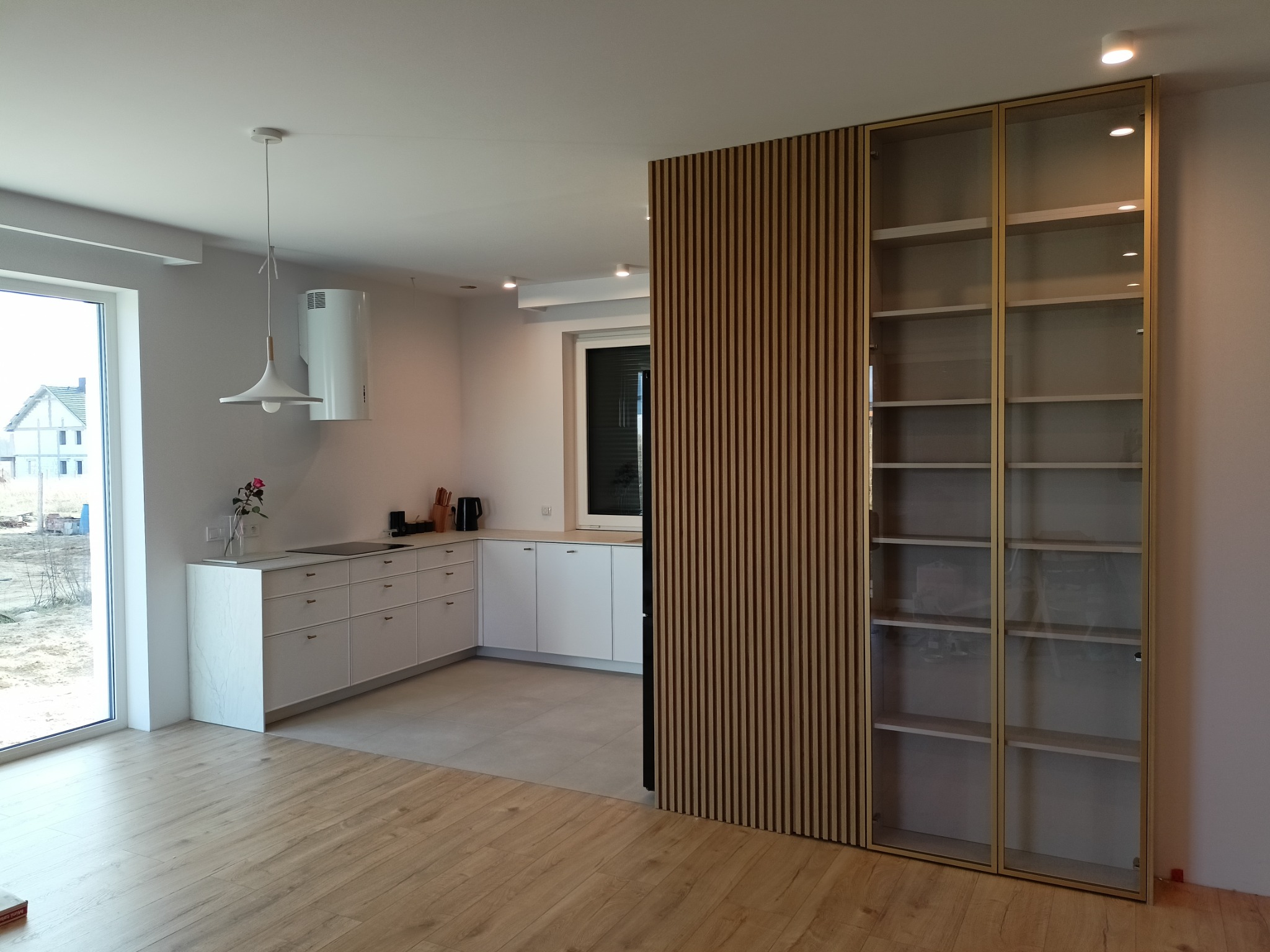 Nowoczesna, jasna kuchnia z białymi szafkami i blatem, oddzielona od salonu drewnianą ścianką działową i przeszkloną szafą z półkami, widok na okno z widokiem na budowę.