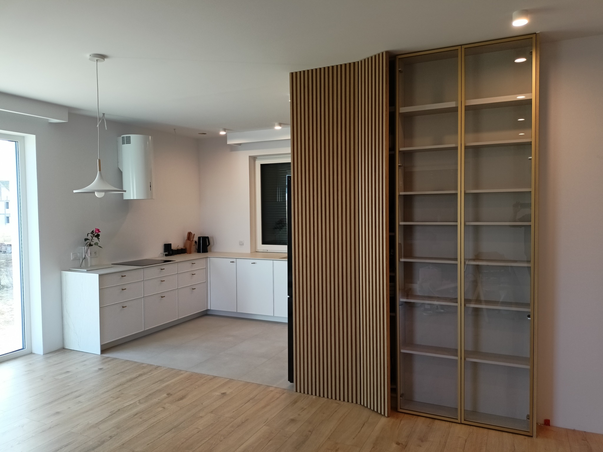 Nowoczesna kuchnia z białymi szafkami, drewnianą ścianką działową z pionowych listew i przeszkloną szafą z półkami, widok na jasne drewniane podłogi.