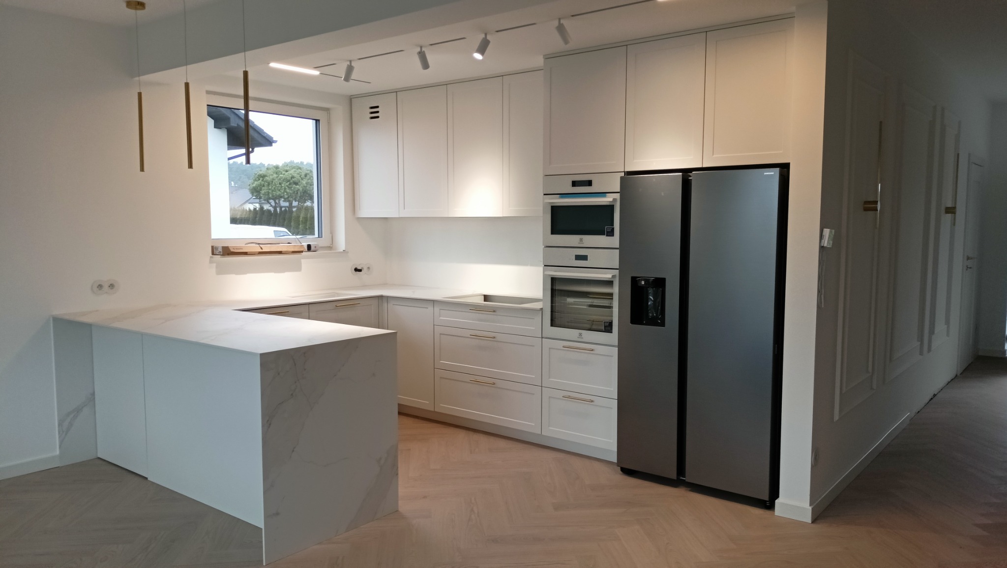 Jasna kuchnia z wyspą i marmurowym blatem. Białe szafki z złotymi uchwytami, sprzęt AGD w zabudowie. Parkiet ułożony w jodełkę. Nowoczesne oświetlenie.