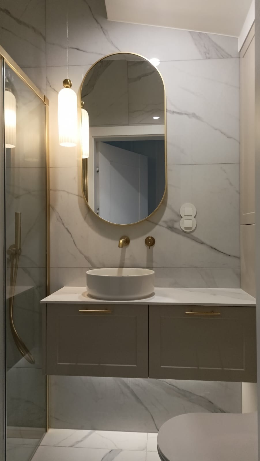 Elegancka łazienka z owalnym lustrem w złotej ramie, umywalką nablatową i szafką pod umywalką z subtelnym oświetleniem LED. Marmurowe płytki na ścianach i podłodze.