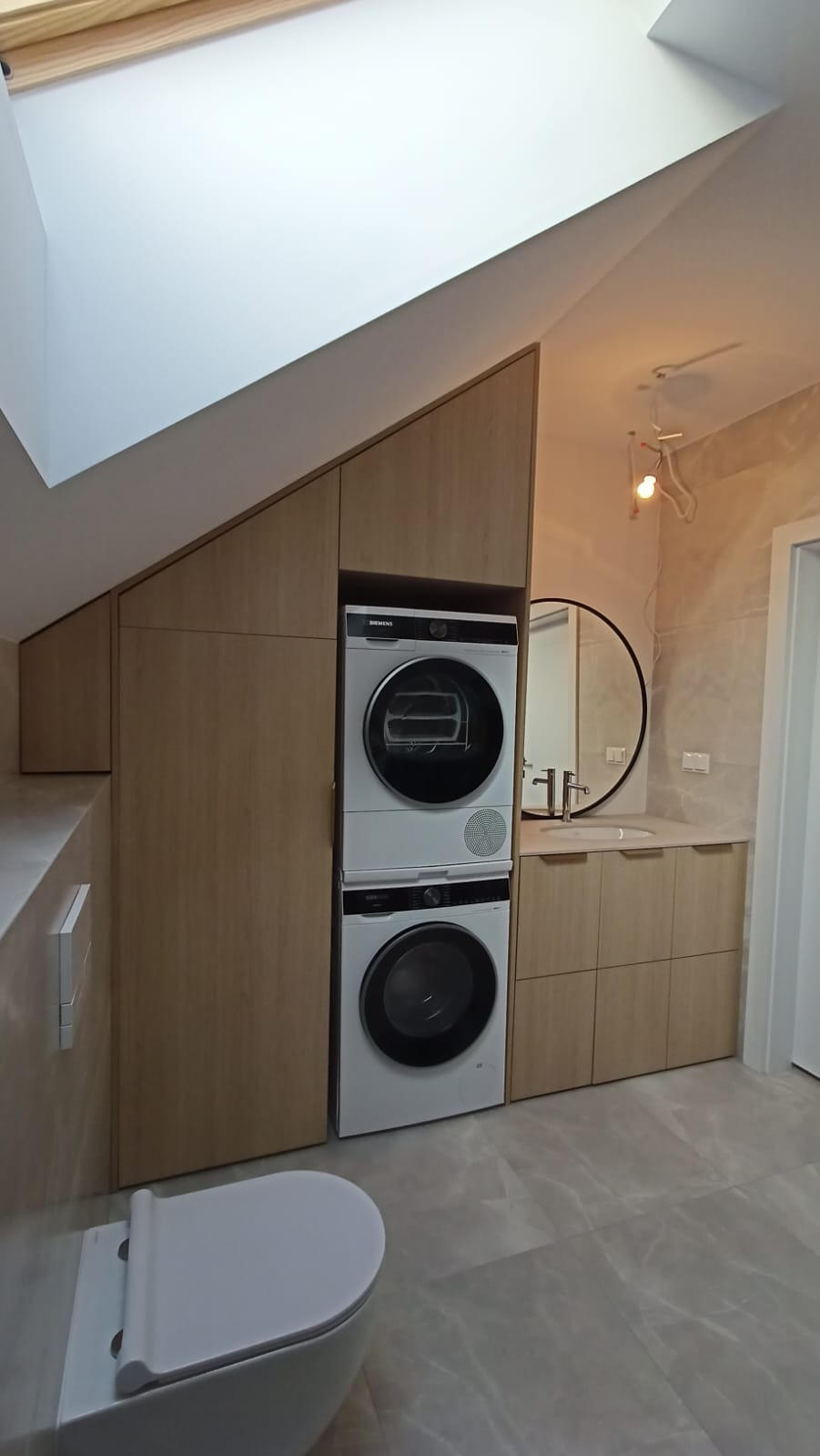 Funkcjonalna zabudowa łazienki na poddaszu w Szczecinie. Jasne drewno, pralka i suszarka w kolumnie, umywalka z szafkami, okrągłe lustro. Wykorzystanie przestrzeni.