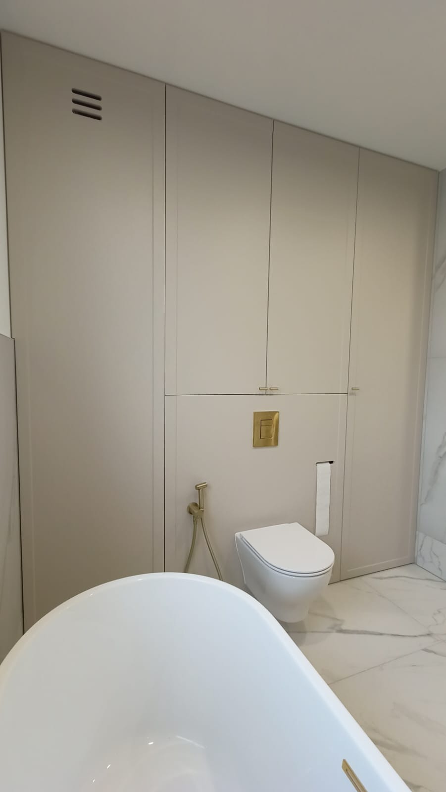 Beżowa szafa wnękowa w łazience z toaletą podwieszaną i złotą armaturą. Widoczny fragment białej wanny i marmurowa podłoga. Minimalistyczny design.