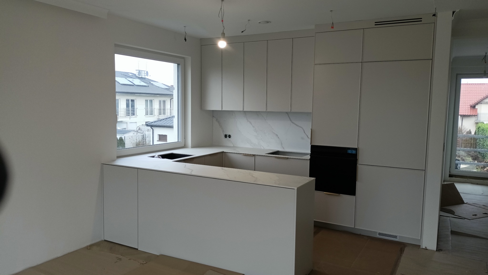 Nowoczesna, jasna kuchnia z wyspą i marmurowym blatem. Minimalistyczne szafki bez uchwytów, okno z widokiem na budynek. Oświetlenie punktowe podkreśla detale.