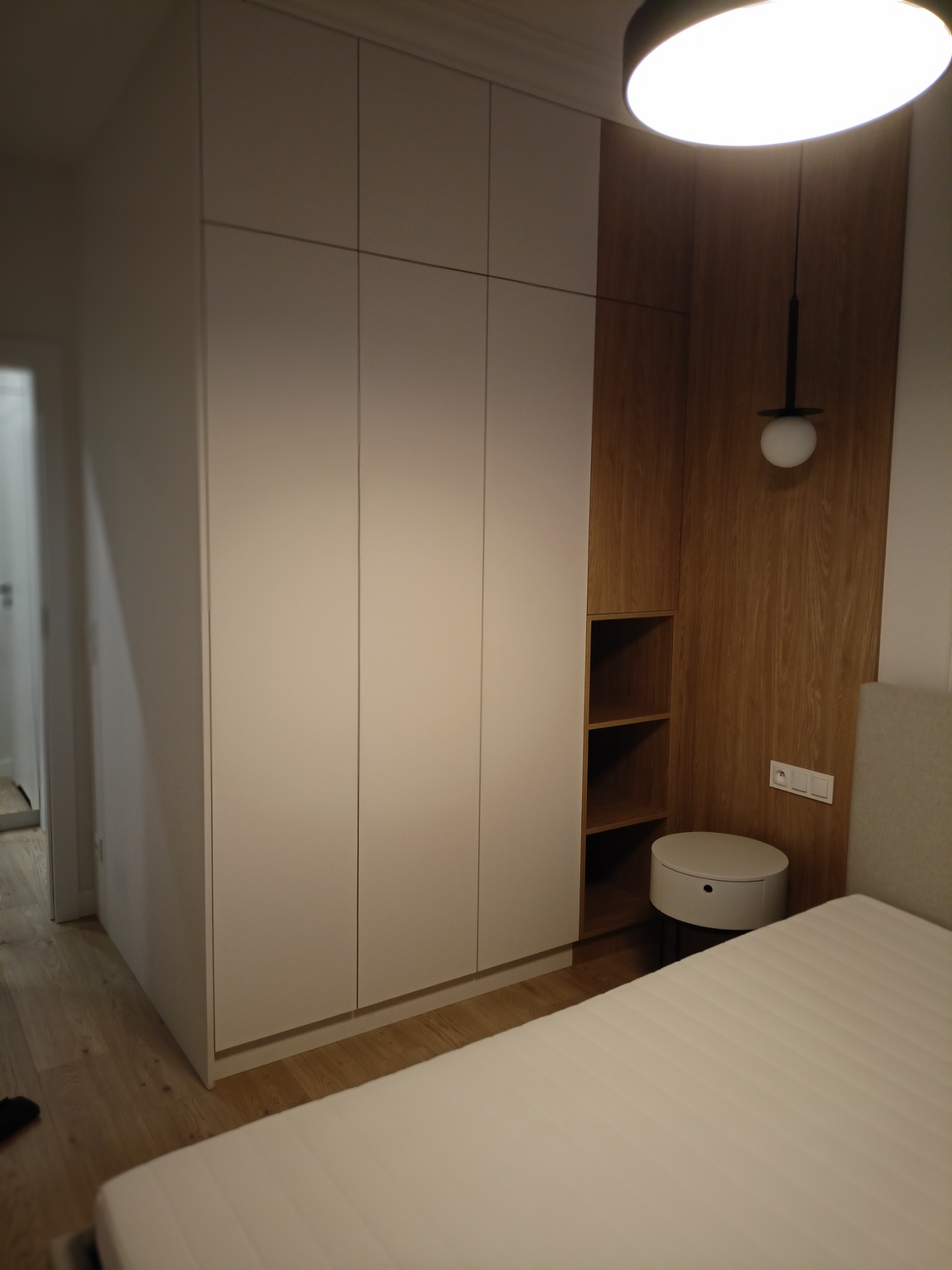 Biała szafa wnękowa z drewnianym panelem i półkami w sypialni, oświetlona lampą sufitową. Minimalistyczny design, jasne wnętrze, widoczny fragment łóżka.