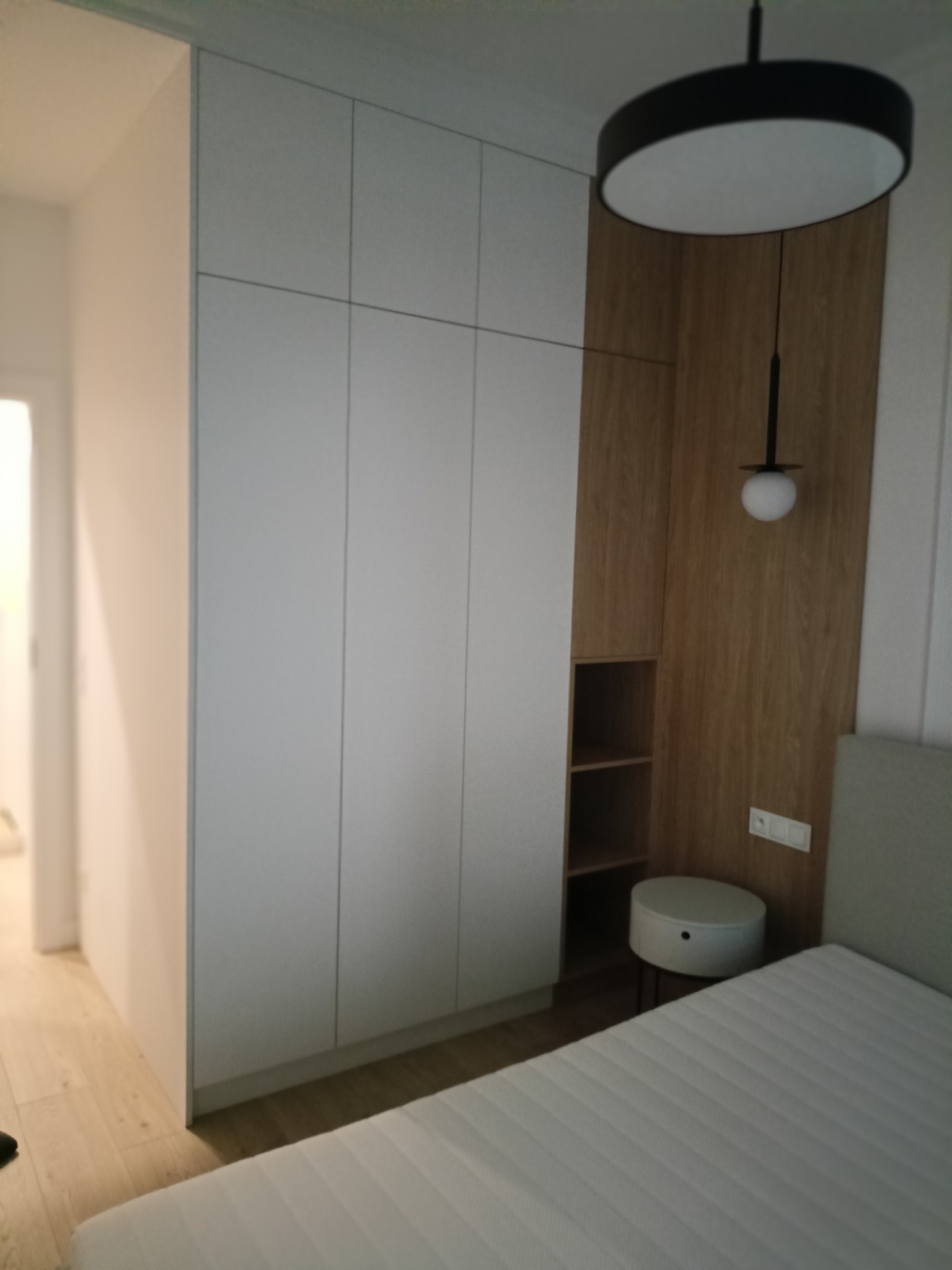 Biała szafa wnękowa z drewnianymi elementami, oświetleniem i szafką nocną w nowoczesnej sypialni. Minimalistyczny design, jasne kolory, funkcjonalne rozwiązanie do przechowywania.