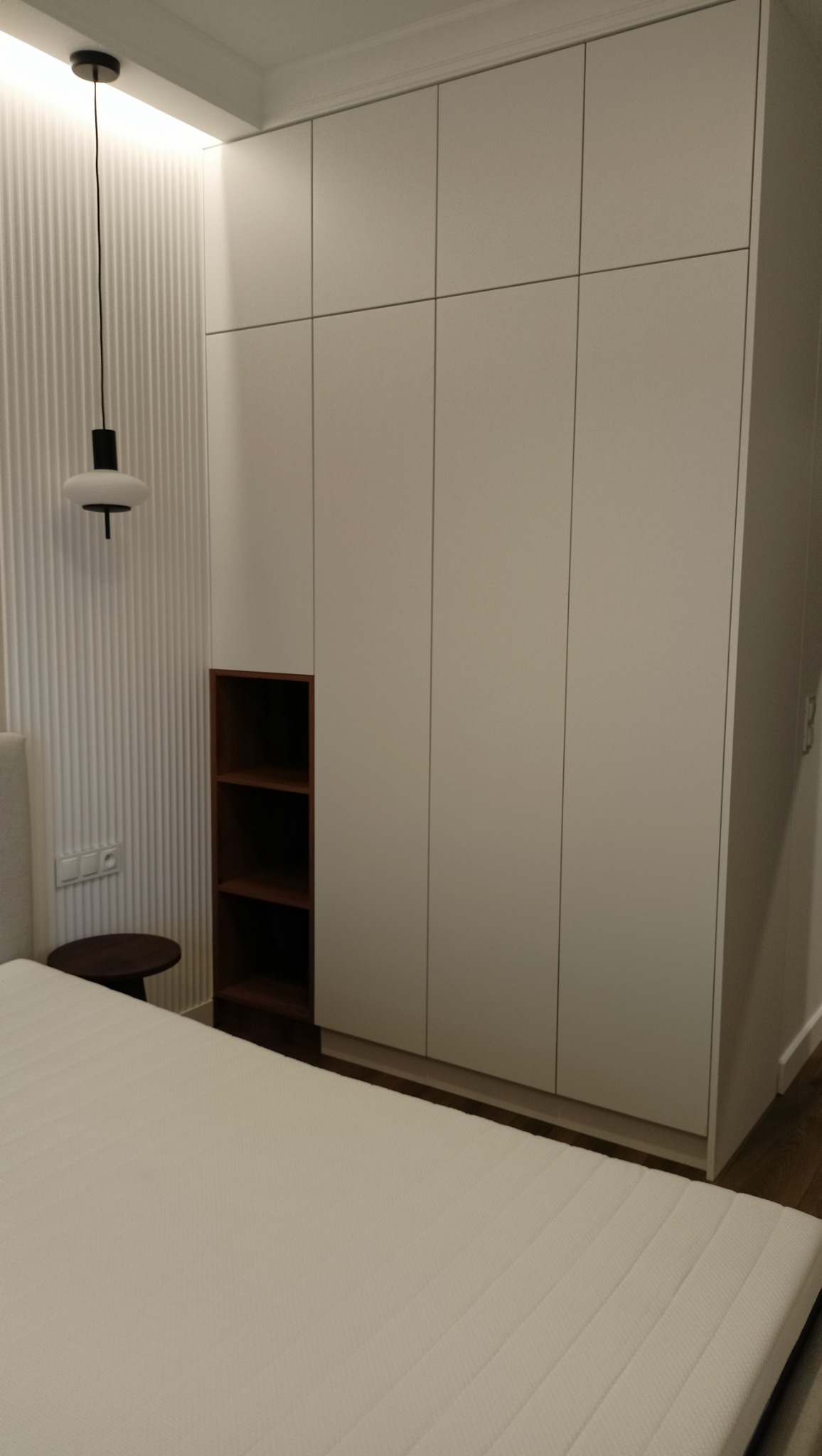 Biała szafa wnękowa z ciemną wnęką na książki, wkomponowana w ścianę sypialni z łóżkiem i lampą wiszącą. Minimalistyczny design, gładkie fronty.