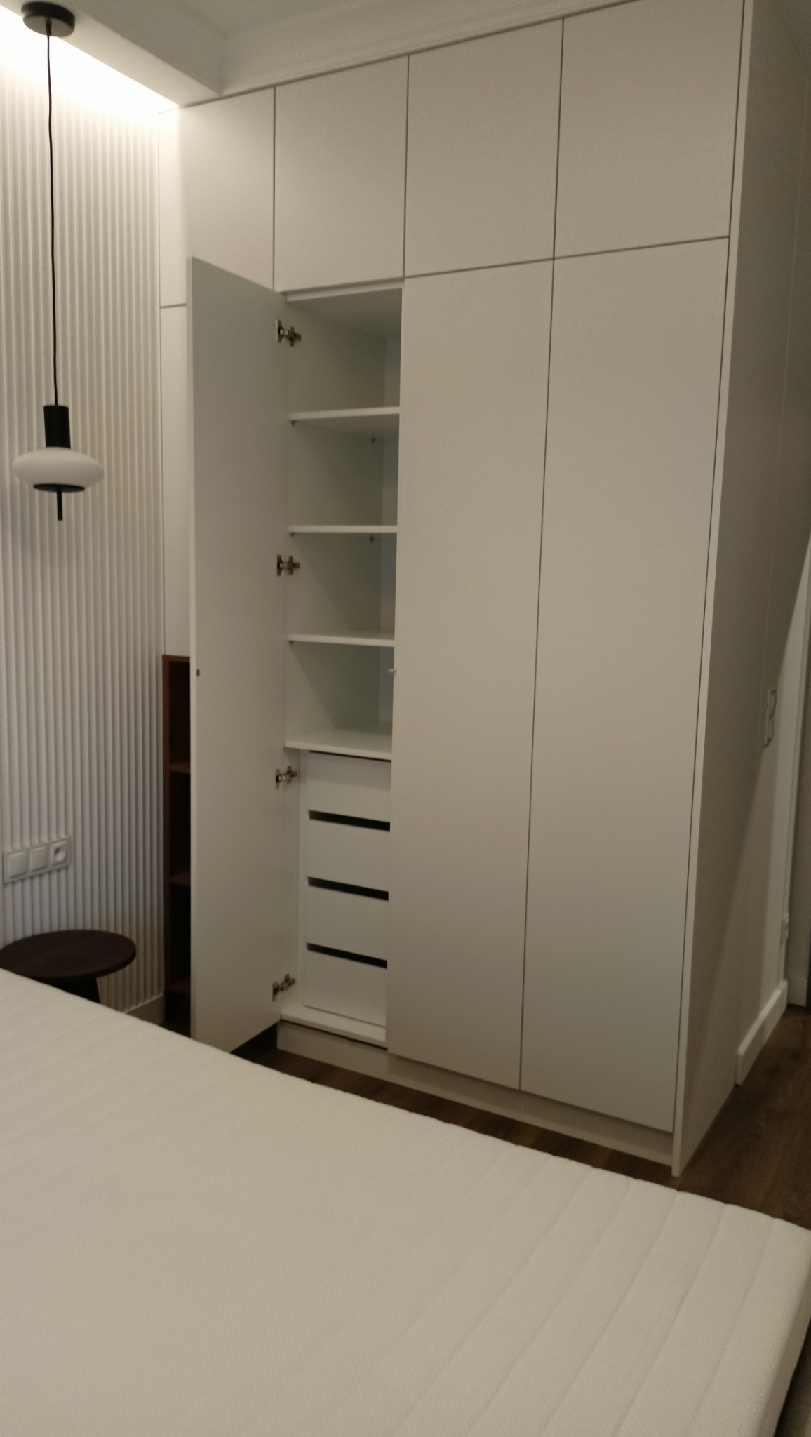 Biała szafa wnękowa z otwartymi drzwiami, widoczne półki i szuflady. Minimalistyczny design, oświetlenie LED w tle. Część łóżka widoczna na pierwszym planie.