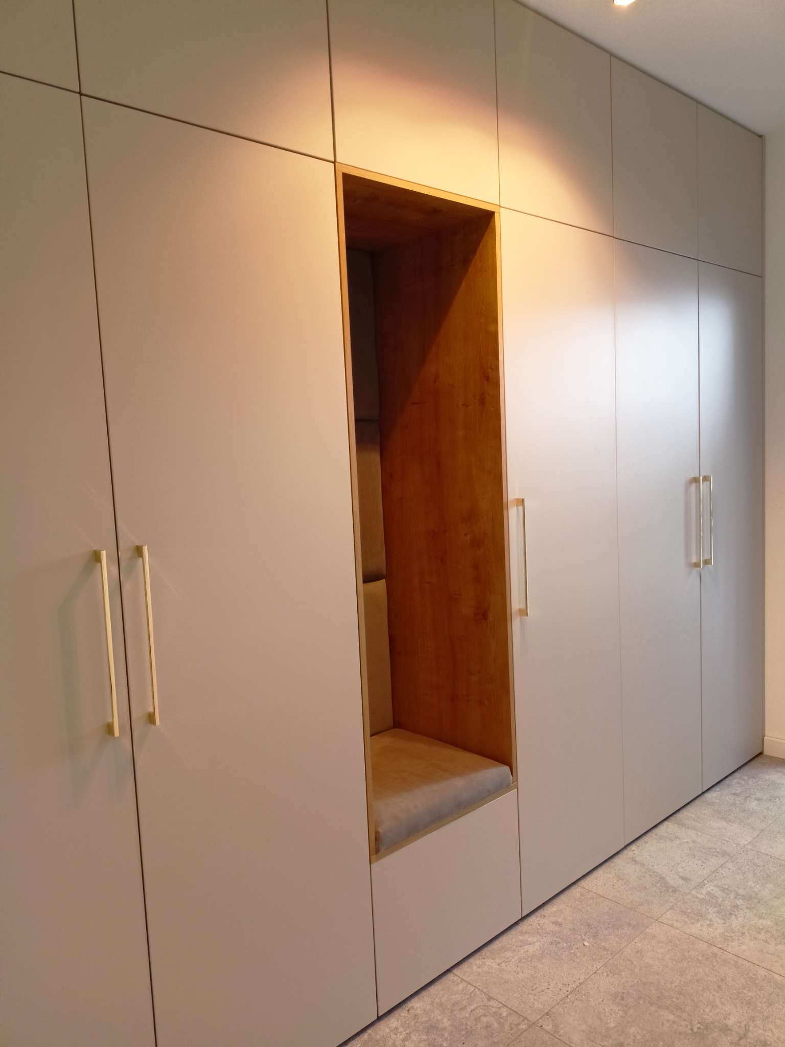 Pojemna szafa w zabudowie z wnęką do siedzenia wykończoną drewnem i tapicerowanym siedziskiem, minimalistyczne złote uchwyty, szare fronty.