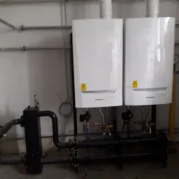 Dwa białe, wiszące piece gazowe zamontowane obok siebie, z czarnymi rurami i zaworami, w surowym, pomalowanym na biało pomieszczeniu z widocznymi rurami instalacyjnymi.