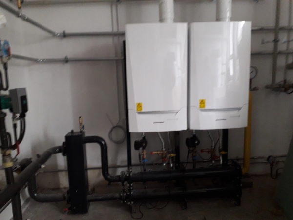 Dwa białe, wiszące piece gazowe zamontowane obok siebie, z czarnymi rurami i zaworami, w surowym, pomalowanym na biało pomieszczeniu z widocznymi rurami instalacyjnymi.