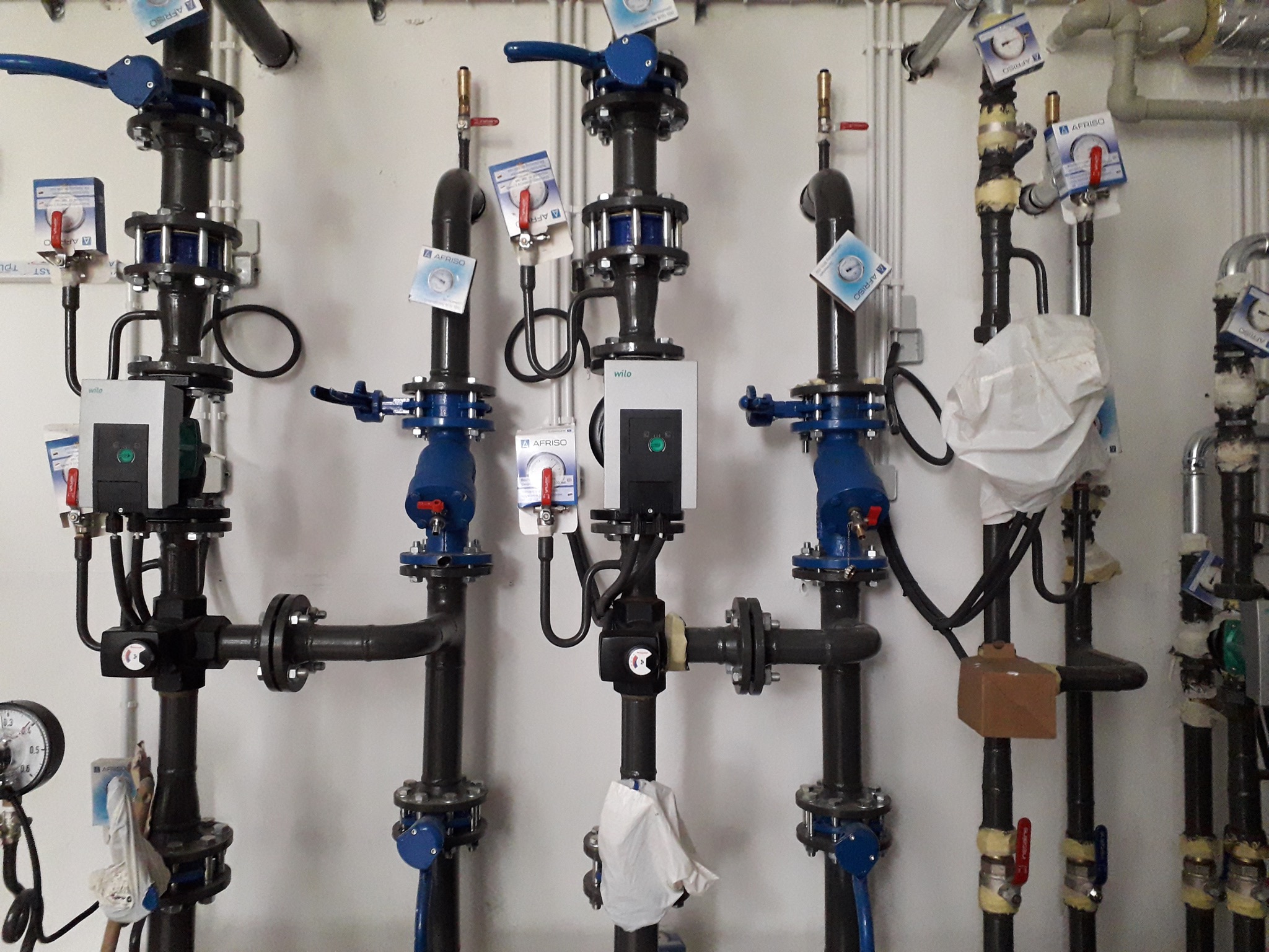 Szereg pionowych rur instalacyjnych z zaworami, manometrami i pompami obiegowymi, widoczne etykiety AFRISO i Wilo, fragment instalacji w kotłowni lub węźle cieplnym.