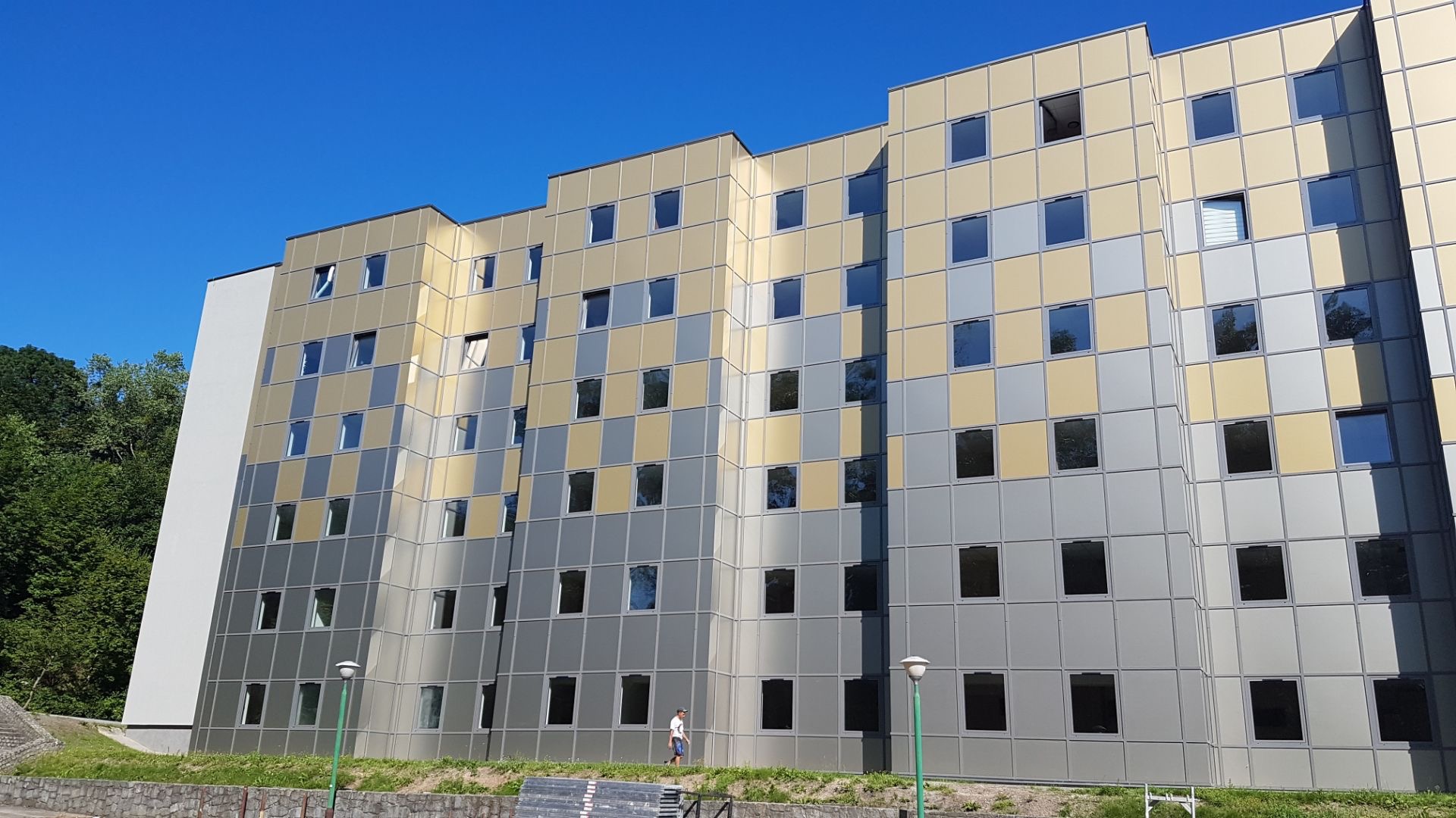 Elewacja budynku z panelami w odcieniach szarości i beżu, regularny układ okien, widoczny fragment zieleni i błękitne niebo.