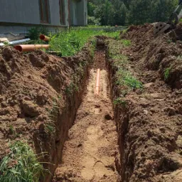 Wykopany rów w ziemi z pomarańczową rurą na dnie, widoczny fragment domu i drzewa w tle, świeżo wykopana ziemia po bokach rowu.