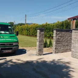 Świeżo postawiona brama wjazdowa z szarej kostki łupanej, z zielonym busem firmy brukarsko-ogrodniczej w tle, na podjeździe z kostki brukowej.