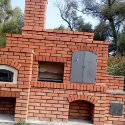 Murowany z czerwonej cegły piec zewnętrzny z metalowymi drzwiczkami i wnękami na drewno, otoczony zielenią.