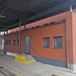 Ceglana elewacja budynku przemysłowego z szarymi oknami i drzwiami, widoczne żółte schody wejściowe i zadaszenie konstrukcji stalowej.