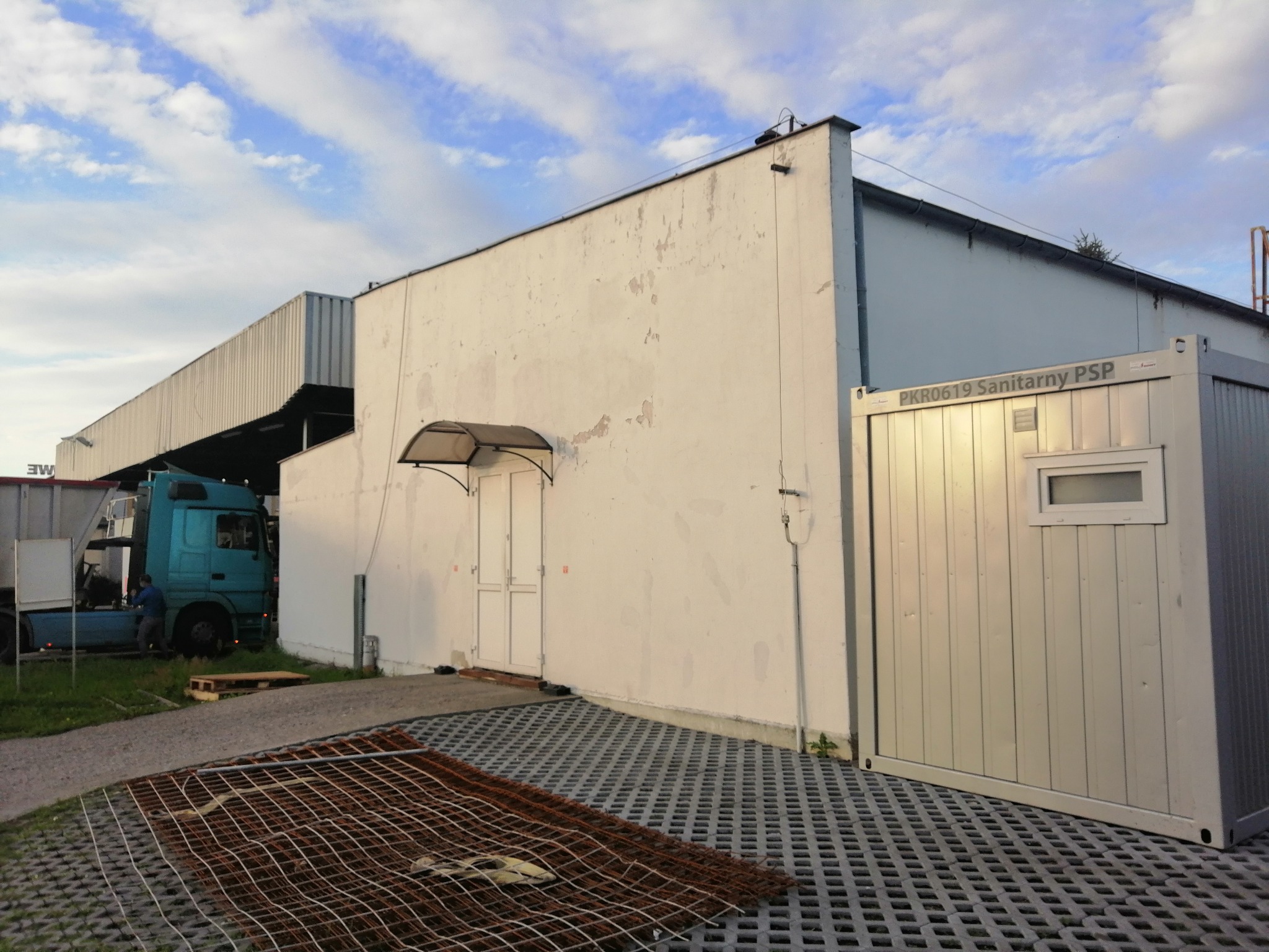 Biała ściana budynku z widocznymi śladami po naprawach, obok kontener sanitarny, na pierwszym planie fragment siatki zbrojeniowej i ażurowa nawierzchnia, w tle zaparkowana ciężarówka.