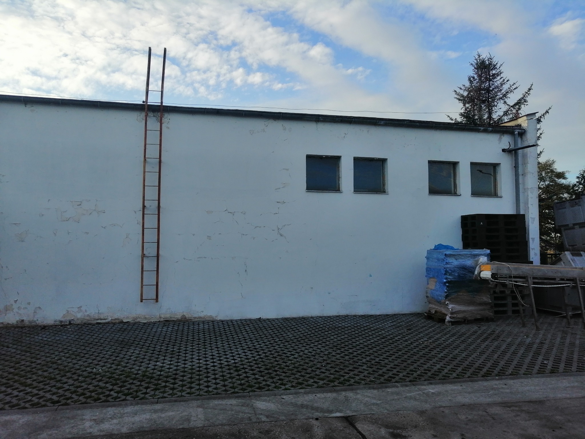 Biała elewacja budynku z trzema małymi oknami, przymocowaną metalową drabiną i popękaną farbą, na dole wyłożona ażurową kostką brukową, z paletami i materiałami budowlanymi w rogu.