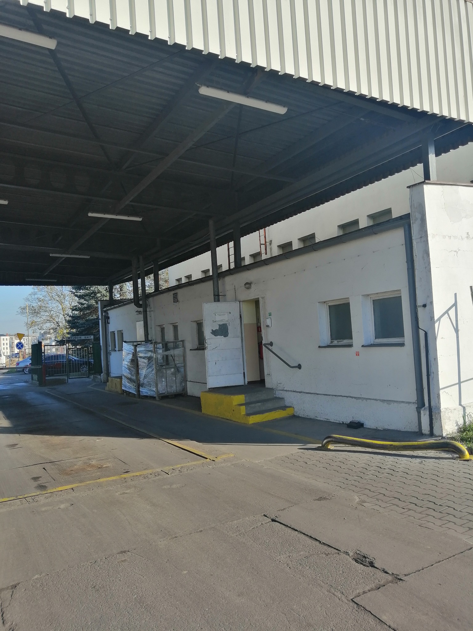 Zewnętrzny widok budynku przemysłowego z rampą załadunkową, żółte elementy zabezpieczające na krawędziach schodów i wzdłuż drogi, zapakowane palety czekają na transport.