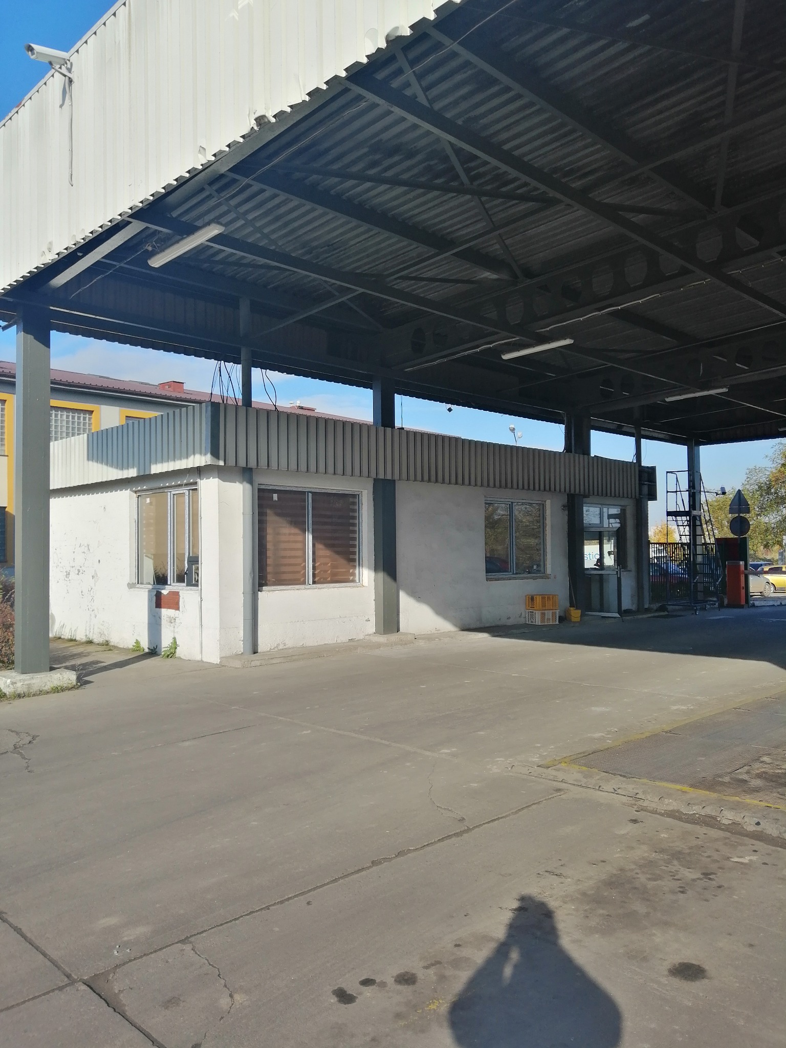 Szara budka strażnicza z płaskim dachem pod zadaszeniem z blachy falistej, widoczne ślady użytkowania na elewacji, żółte skrzynki i wiadro obok wejścia.