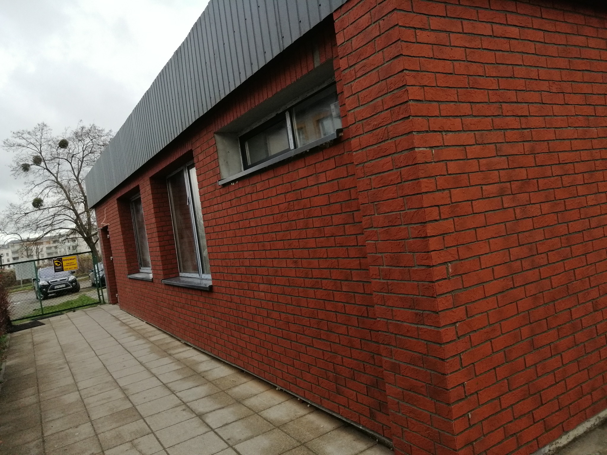 Czerwona ceglana ściana budynku z trzema oknami, szara metalowa blacha na dachu, widok z boku na chodnik z kostki brukowej, drzewo w tle.