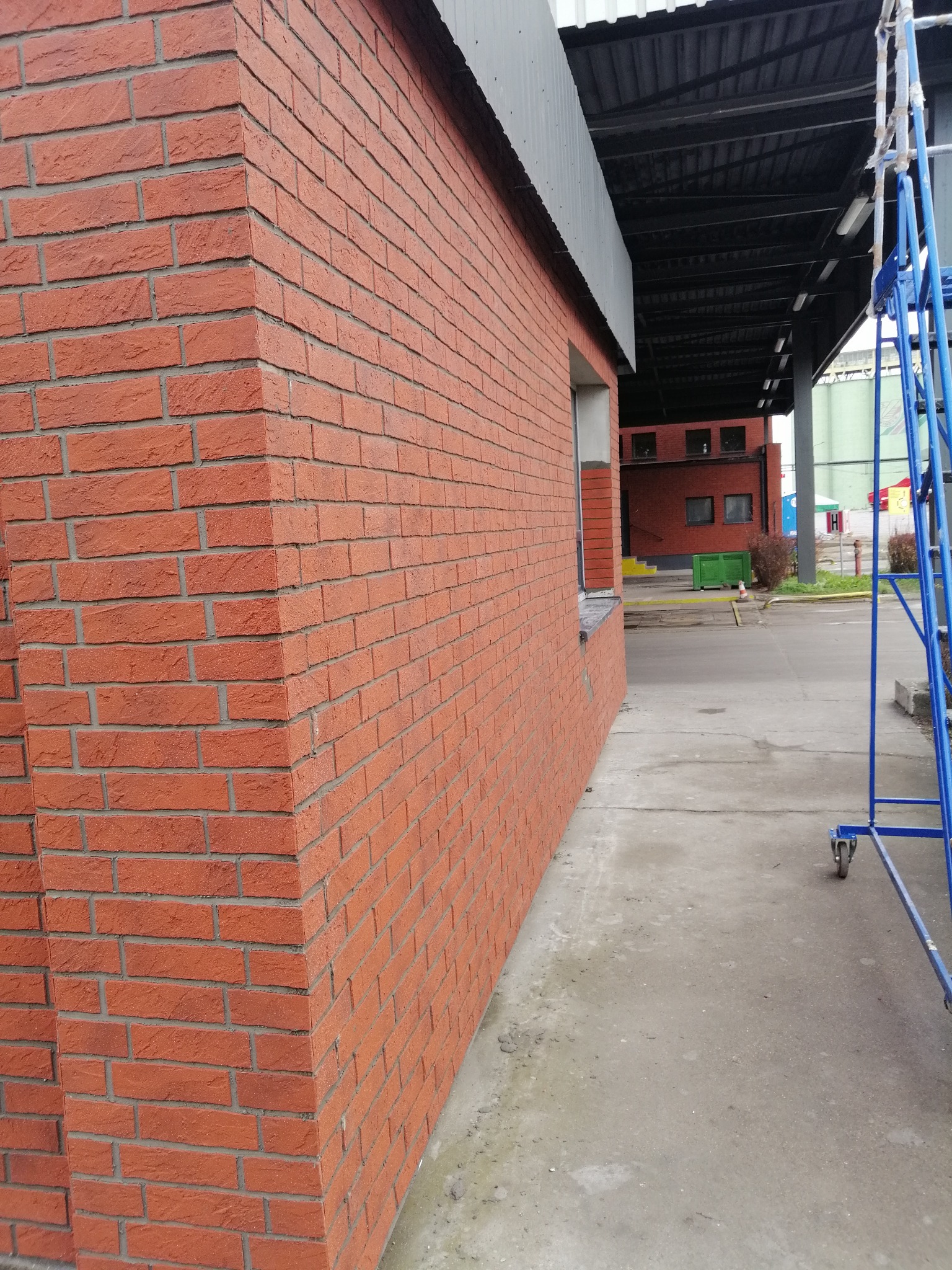 Czerwona ściana z cegły klinkierowej budynku komercyjnego, widok z boku z fragmentem rusztowania, betonowy chodnik i zadaszenie w tle.
