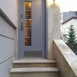 Szare drzwi wejściowe z szybą i metalowymi listwami, zamontowane w kamiennej elewacji budynku w Krakowie, widoczne schody prowadzące do wejścia.