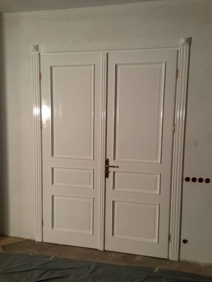 Białe, dwuskrzydłowe drzwi wewnętrzne z dekoracyjnymi listwami, zamontowane w świeżo pomalowanej ścianie, zabezpieczone folią malarską na podłodze.
