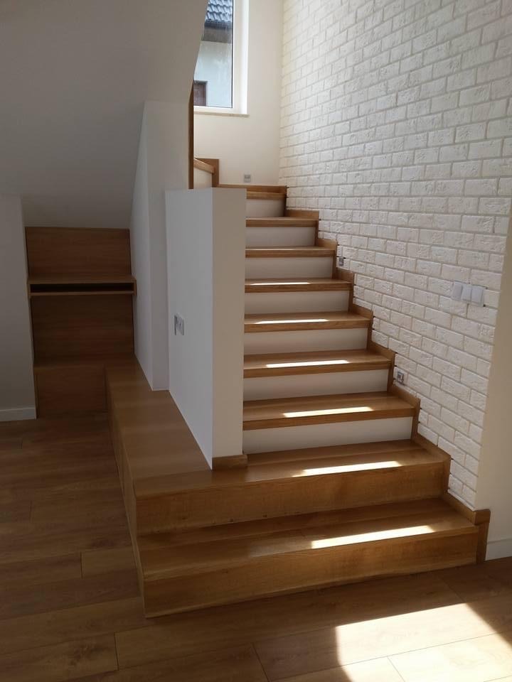 Jasne, drewniane stopnie schodów z białymi podstopnicami prowadzące w górę, oświetlone promieniami słońca padającymi przez okno, z białymi cegłami na ścianie i wbudowaną szafką w przestrzeni...