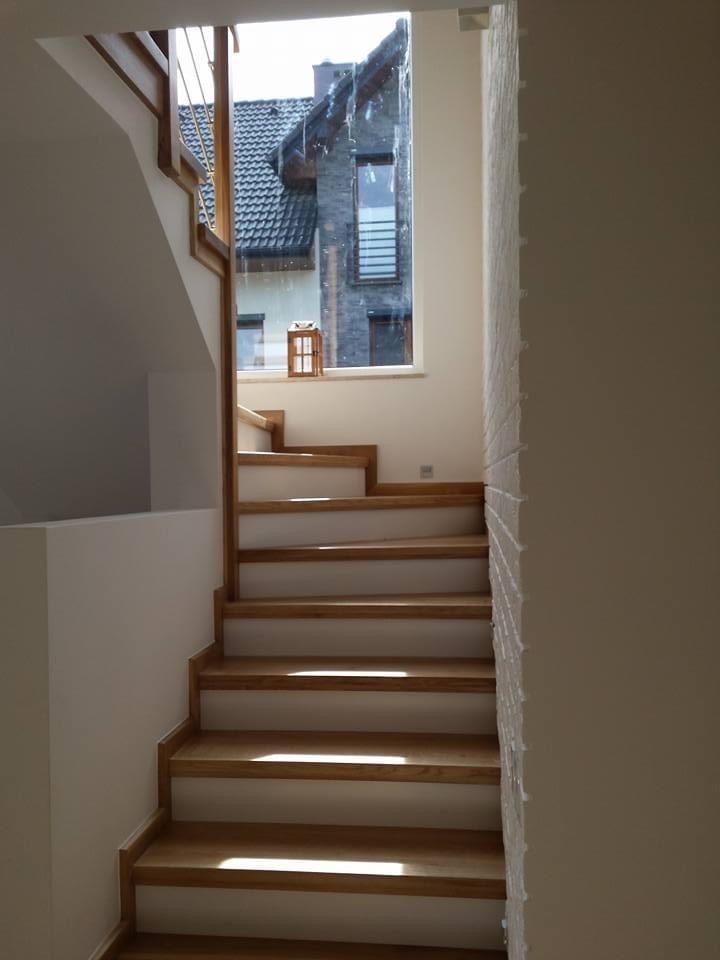 Wewnętrzne schody z jasnego drewna z białymi podstopnicami, oświetlone naturalnym światłem wpadającym przez okno z widokiem na dom z ciemnej cegły.