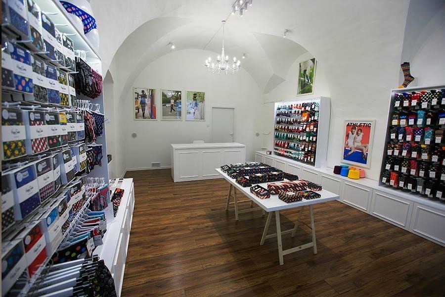 Wnętrze sklepu z białymi ścianami i sklepieniem łukowym prezentuje ekspozycję kolorowych skarpetek w opakowaniach, ustawionych na półkach i na białym stole ekspozycyjnym.