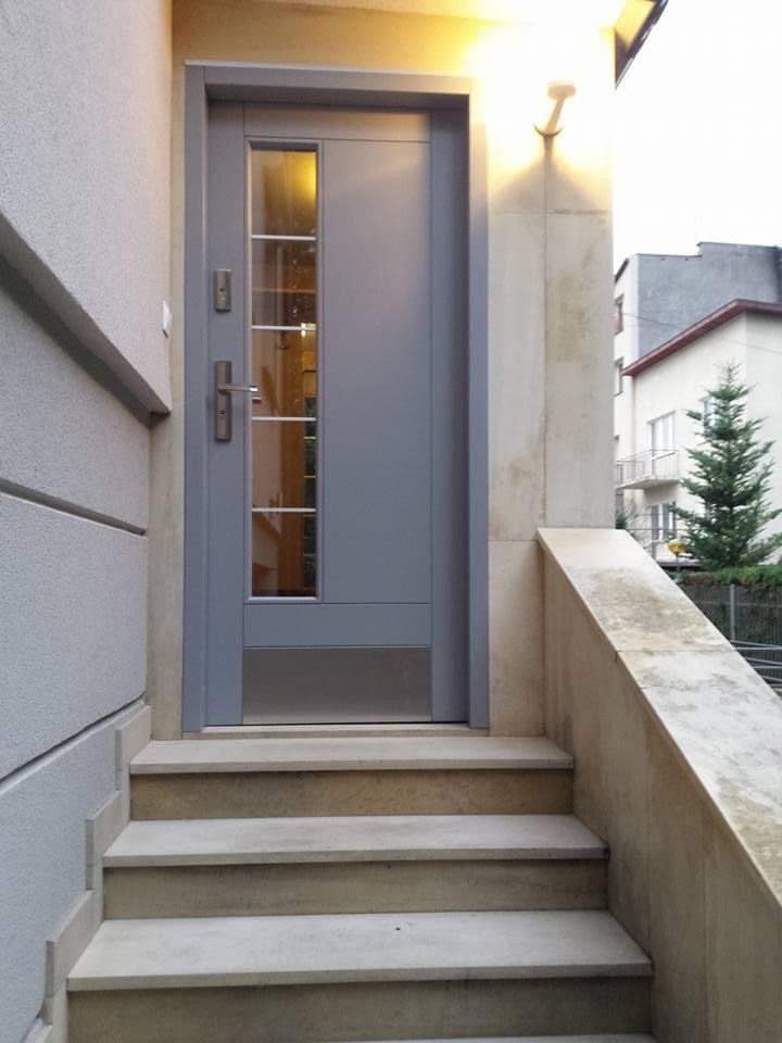 Szare drzwi wejściowe z szybą i metalowymi listwami, zamontowane w kamiennej elewacji budynku w Krakowie, widoczne schody prowadzące do wejścia.