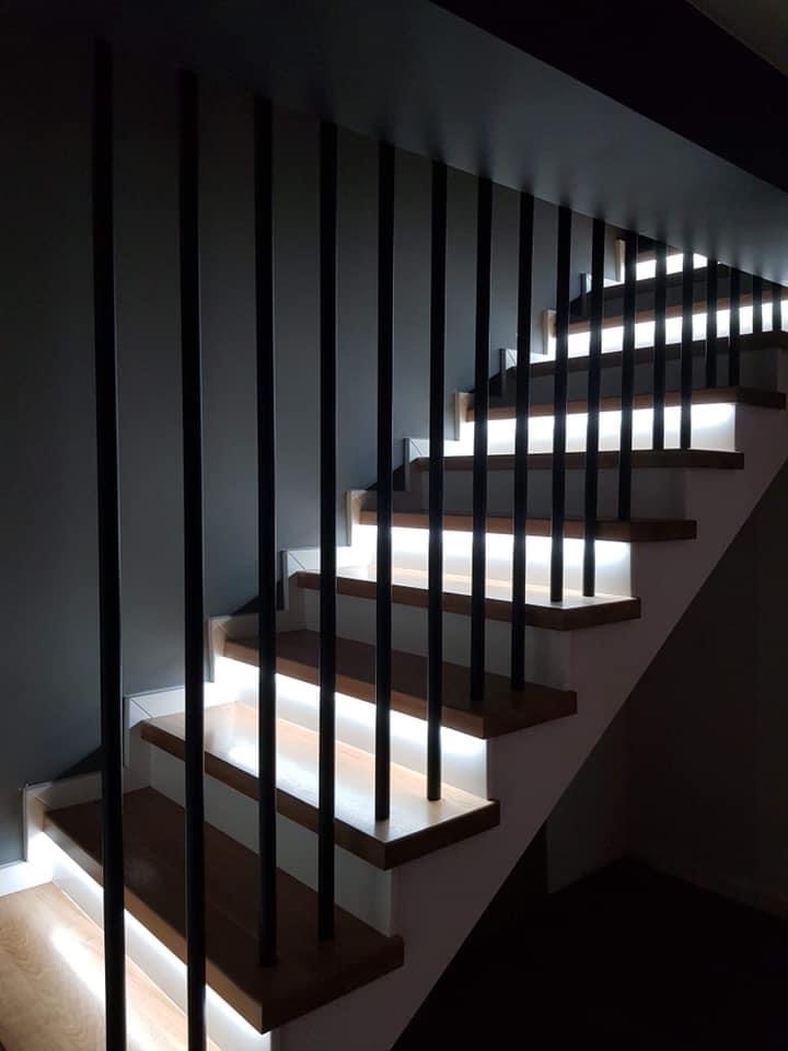 Nowoczesne schody z drewnianymi stopniami, białymi podstopnicami i czarnymi, pionowymi balustradami, podświetlone ledami od spodu.
