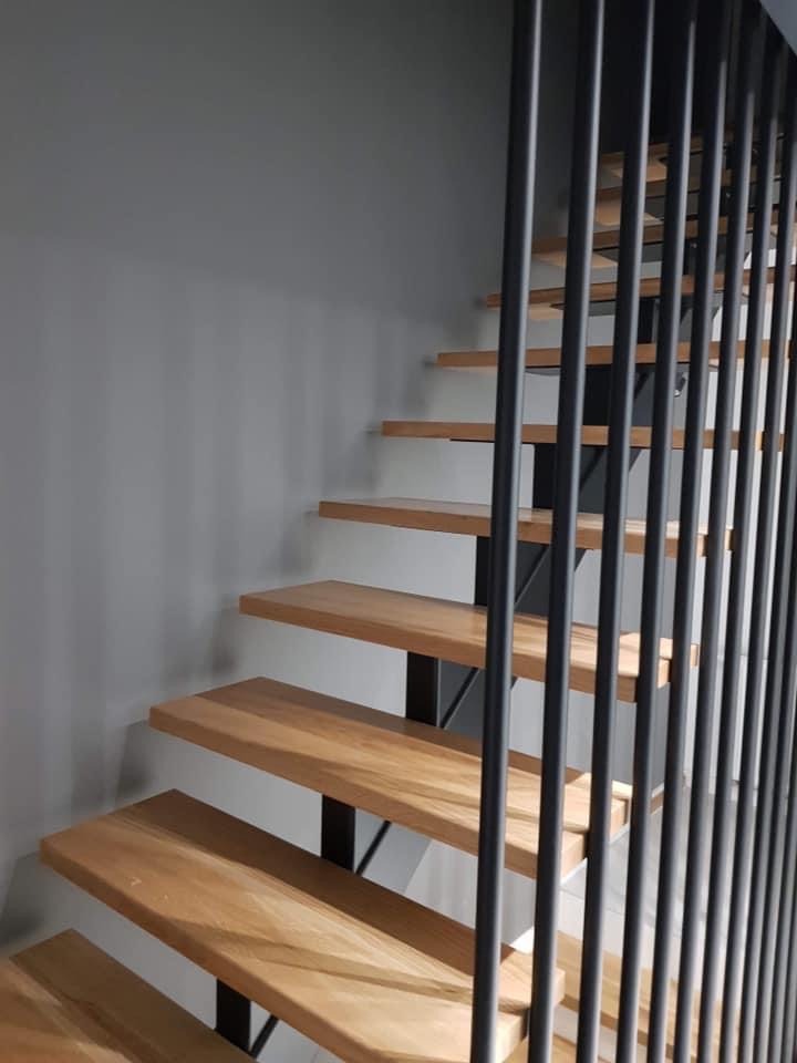 Nowoczesne schody z drewnianymi stopniami i czarną metalową balustradą z pionowych prętów, widok z dołu na tle szarej ściany.