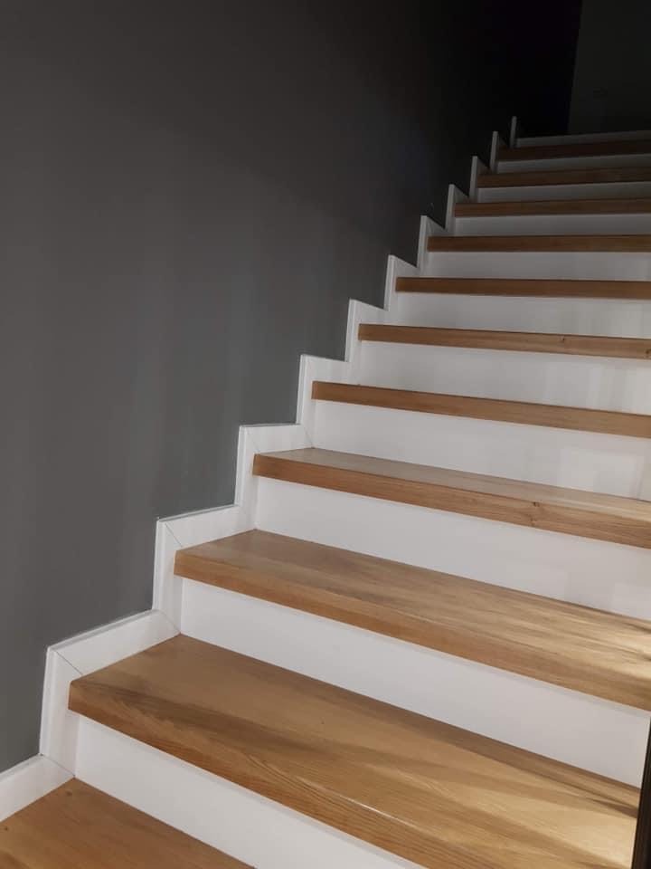Bieg schodów z drewnianymi stopniami i białymi podstopnicami, widok z dołu, szara ściana po lewej stronie.