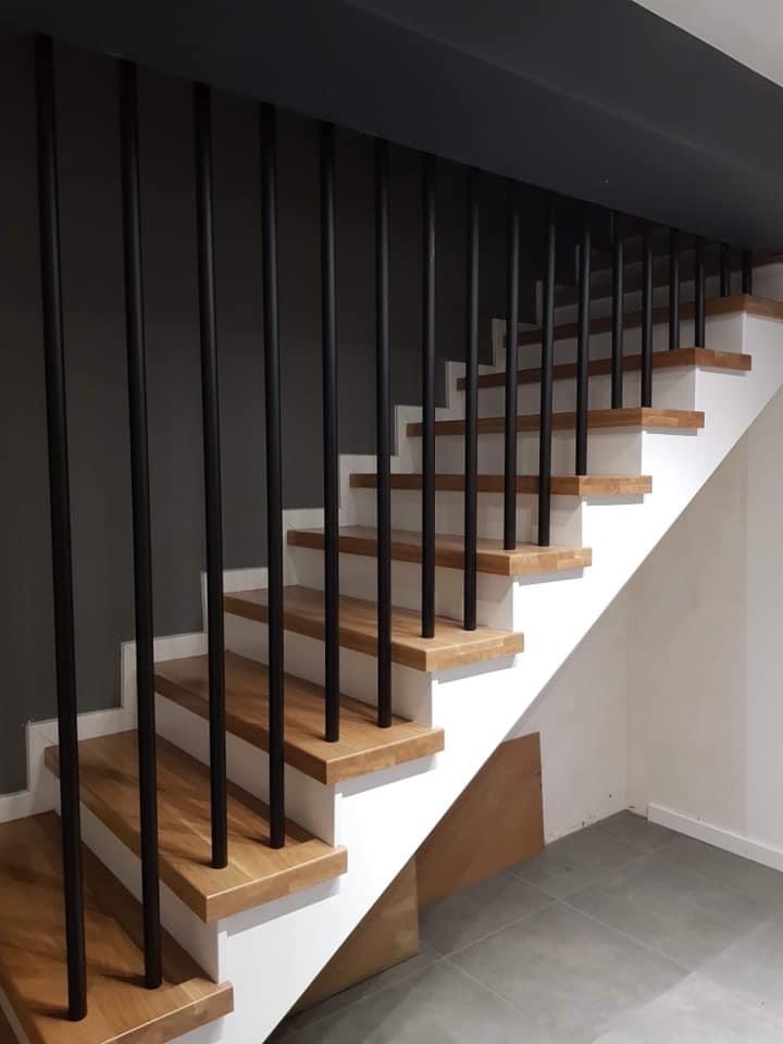 Drewniane schody z białymi podstopnicami i czarnymi, pionowymi balustradami na tle ciemnoszarej ściany i szarej podłogi.