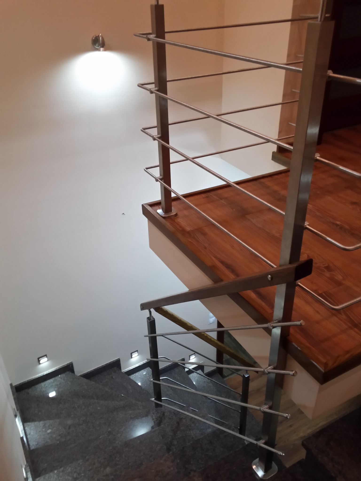 Wewnętrzne schody z ciemnego granitu z podświetleniem LED, kontrastujące z drewnianymi stopniami i nowoczesną balustradą ze stali nierdzewnej, oświetlone punktowym światłem na ścianie.