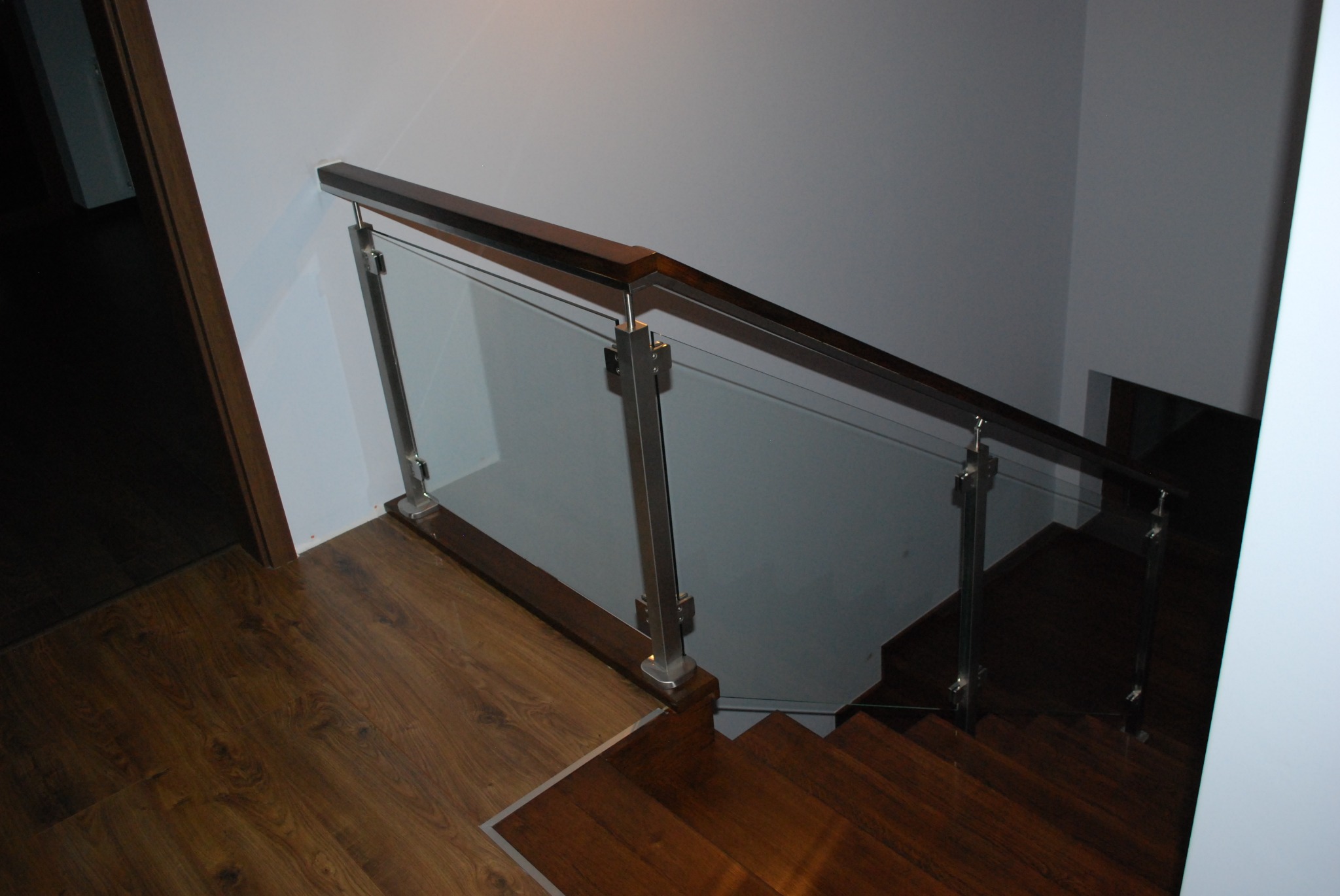 Nowoczesne schody z drewnianymi stopniami i szklaną balustradą z metalowymi słupkami, widoczne z góry, z parkietem w tle.