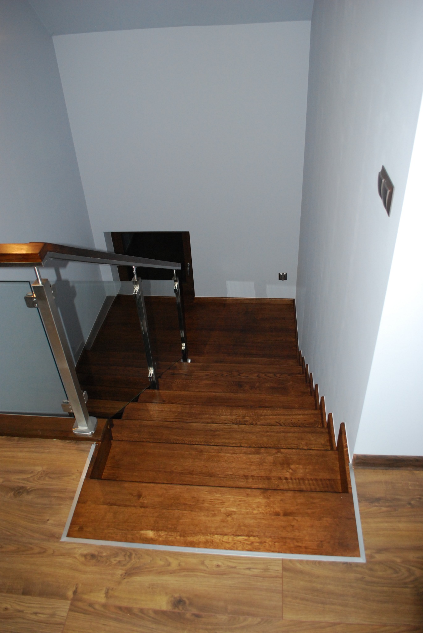 Widok z góry na dębowe stopnie schodów z szklaną balustradą i metalowymi elementami, prowadzące w dół do ciemnego pomieszczenia, z widocznymi listwami przypodłogowymi i przełącznikiem na ścianie.
