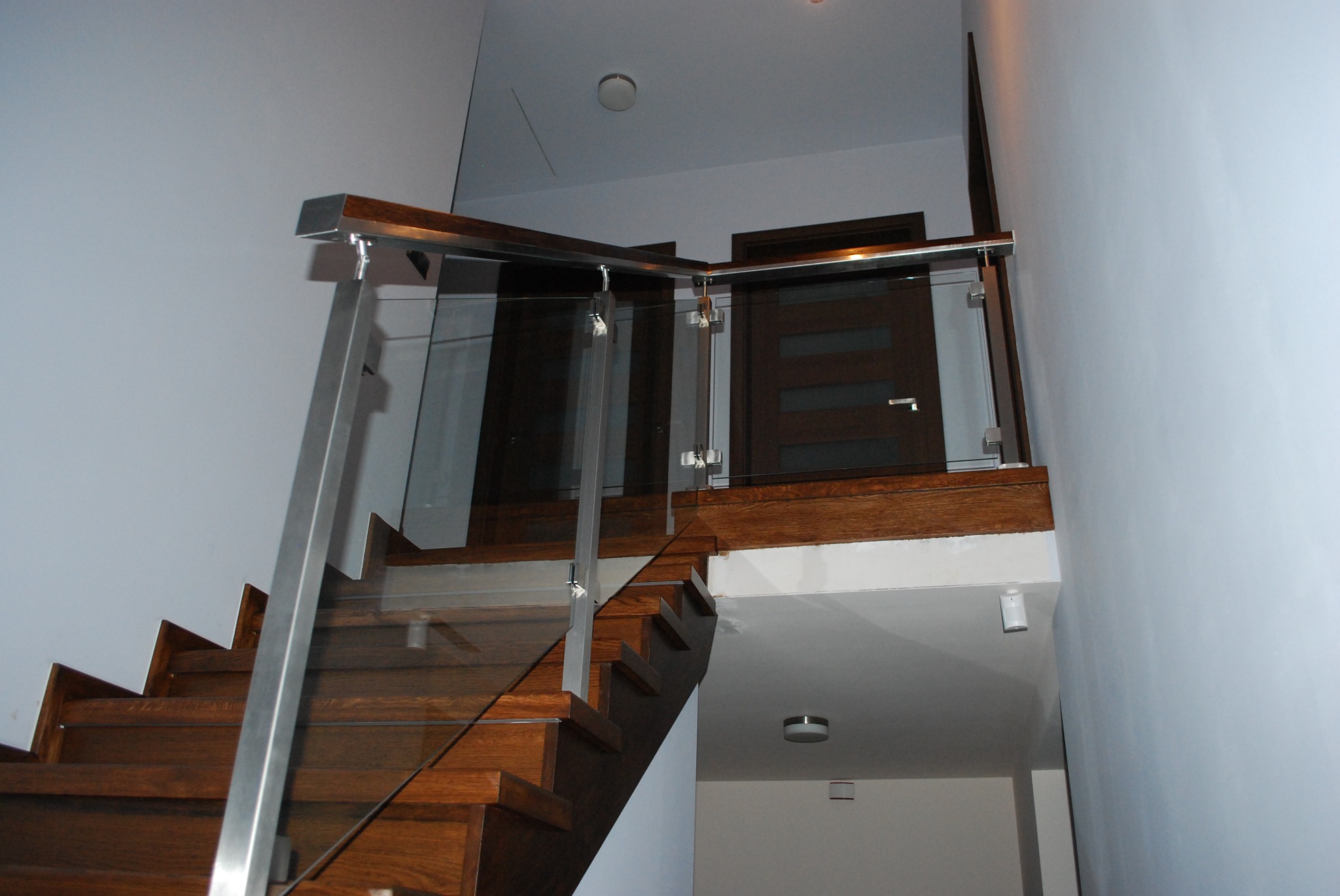 Nowoczesne schody drewniane z balustradą ze szkła i stali nierdzewnej, prowadzące na antresolę z ciemnymi drzwiami.
