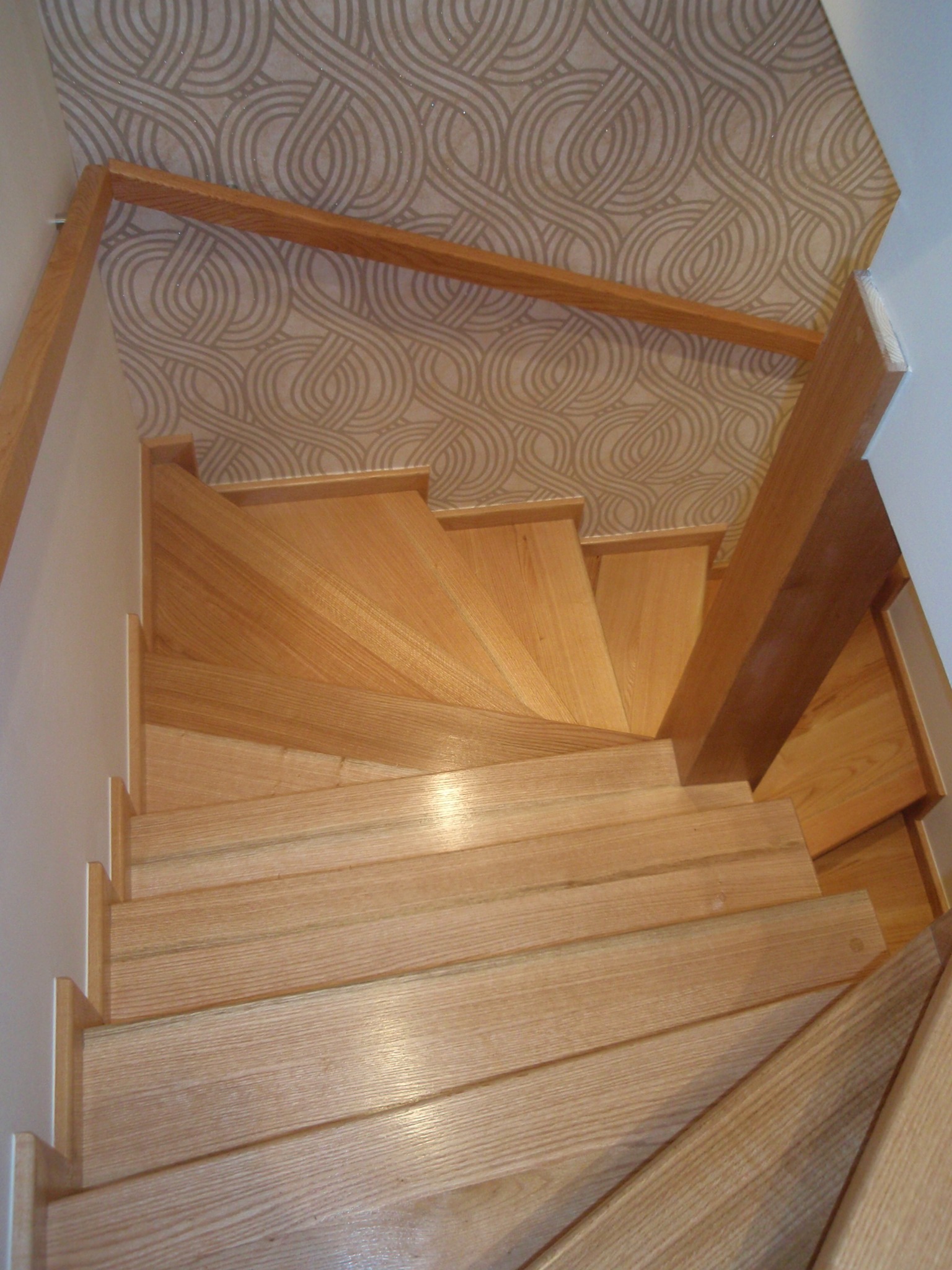 Widok z góry na jasne, drewniane schody skręcające w prawo, z beżową tapetą w geometryczne wzory na ścianie w tle.