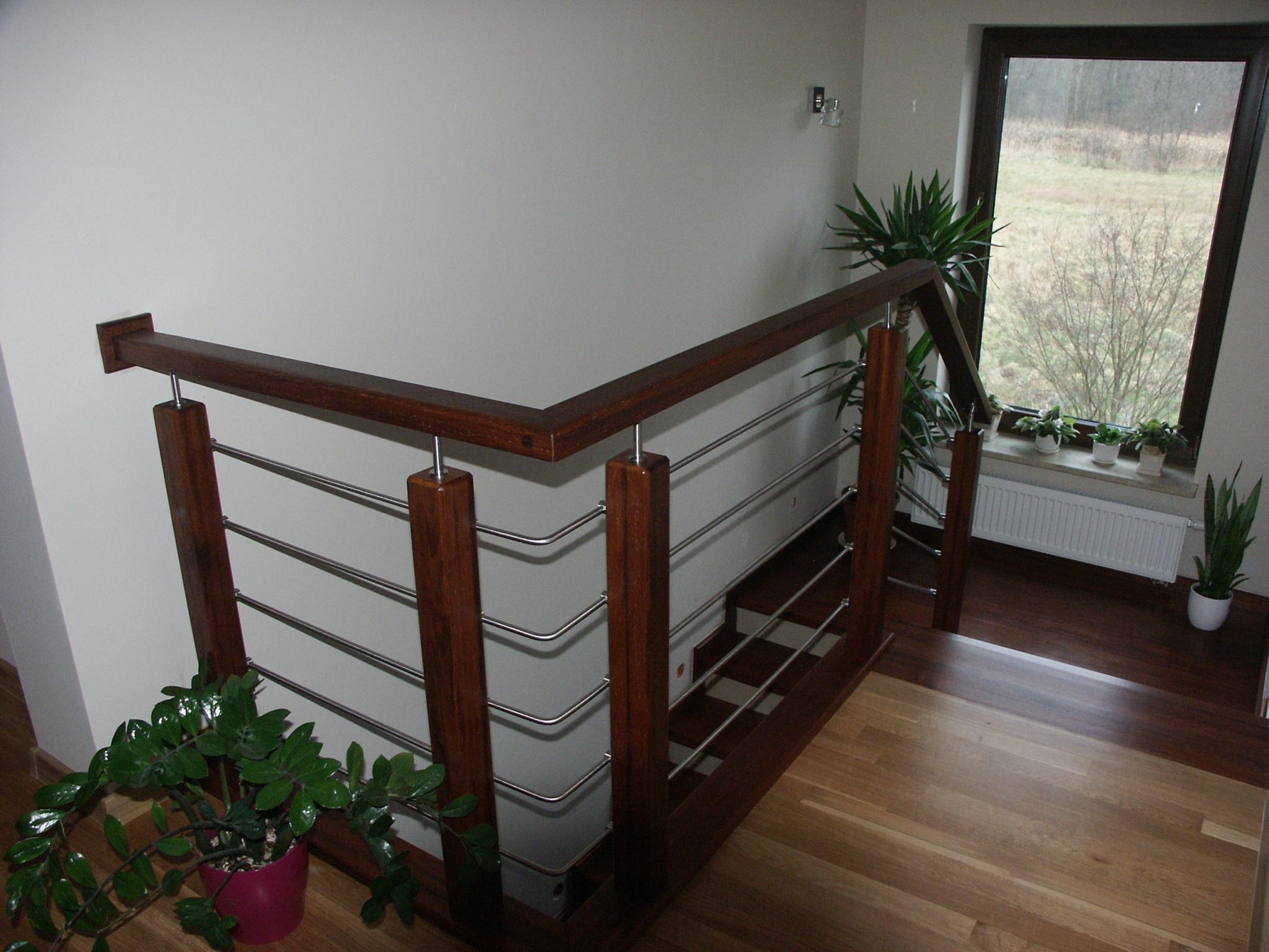 Wnętrze domu z drewnianymi schodami z metalowymi balustradami, widok z góry na podest, z roślinami doniczkowymi i oknem z widokiem na zewnątrz.