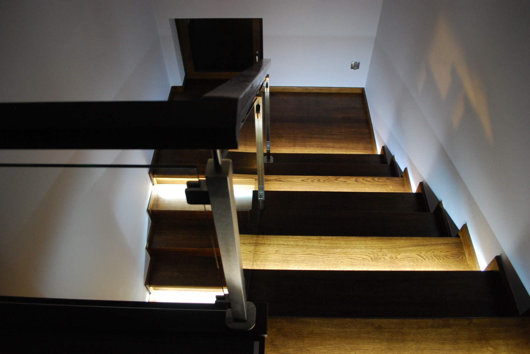 Nowoczesne schody z drewnianymi stopniami w odcieniach jasnego i ciemnego brązu, z metalowymi i szklanymi elementami balustrady, oświetlone od spodu stopnic.